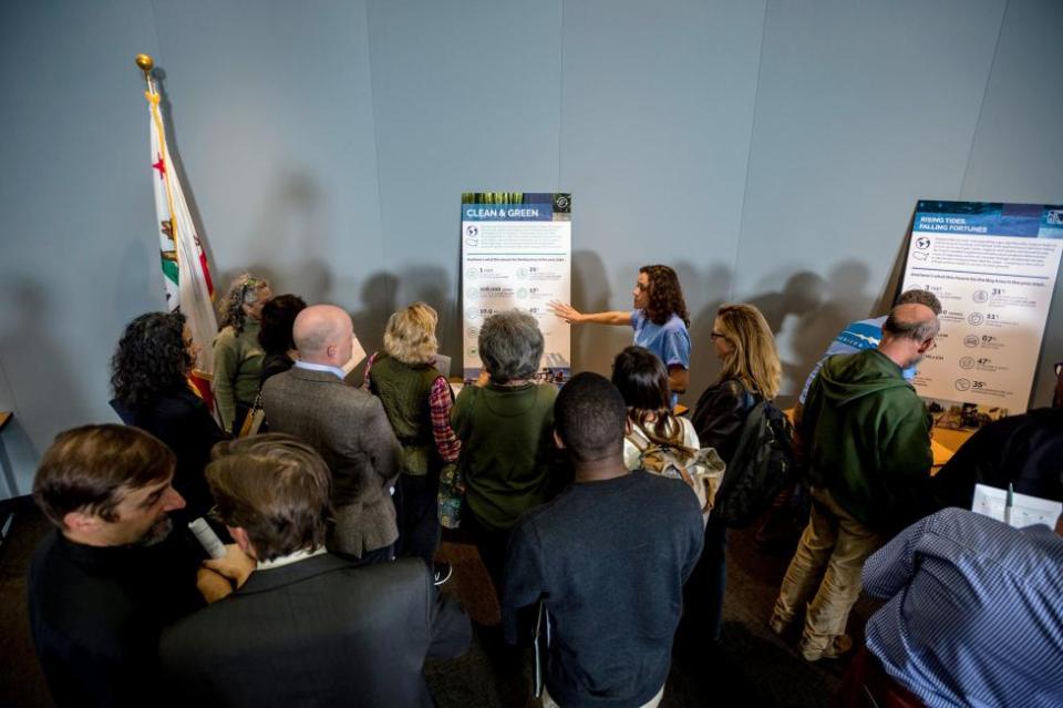 Staff stand near display boards and answer questions from a crowd of attendees at a public workshop.