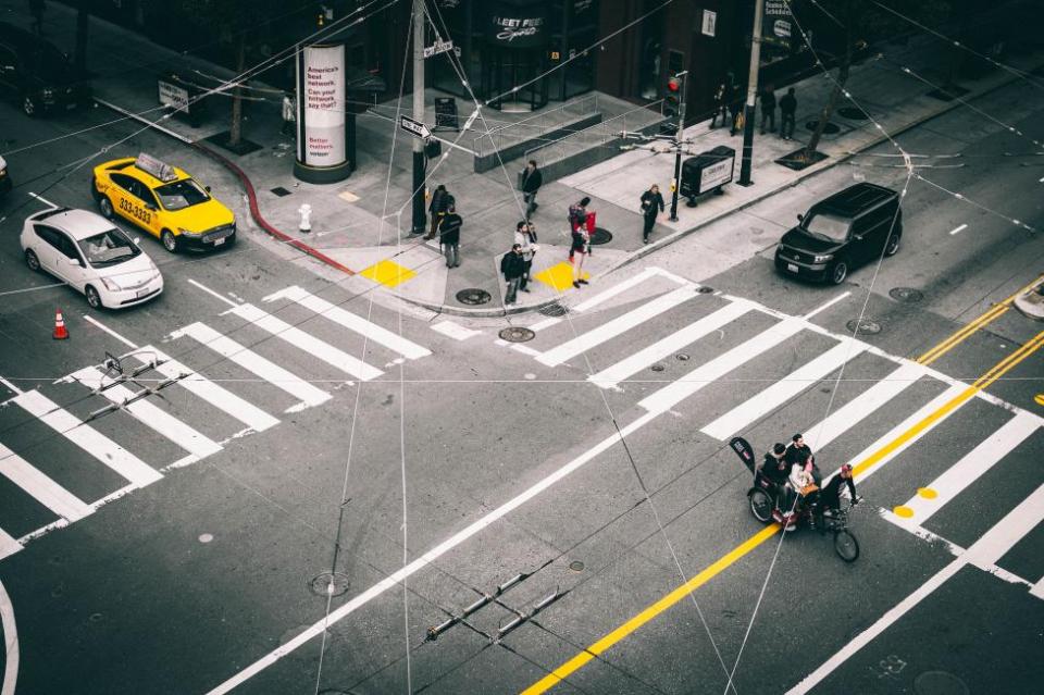 Pedestrians wait at the crosswalk, a pedi-cab crosses the intersection, and cars and taxis wait. The view is seen from above through criss-crossing Muni overhead wires.
