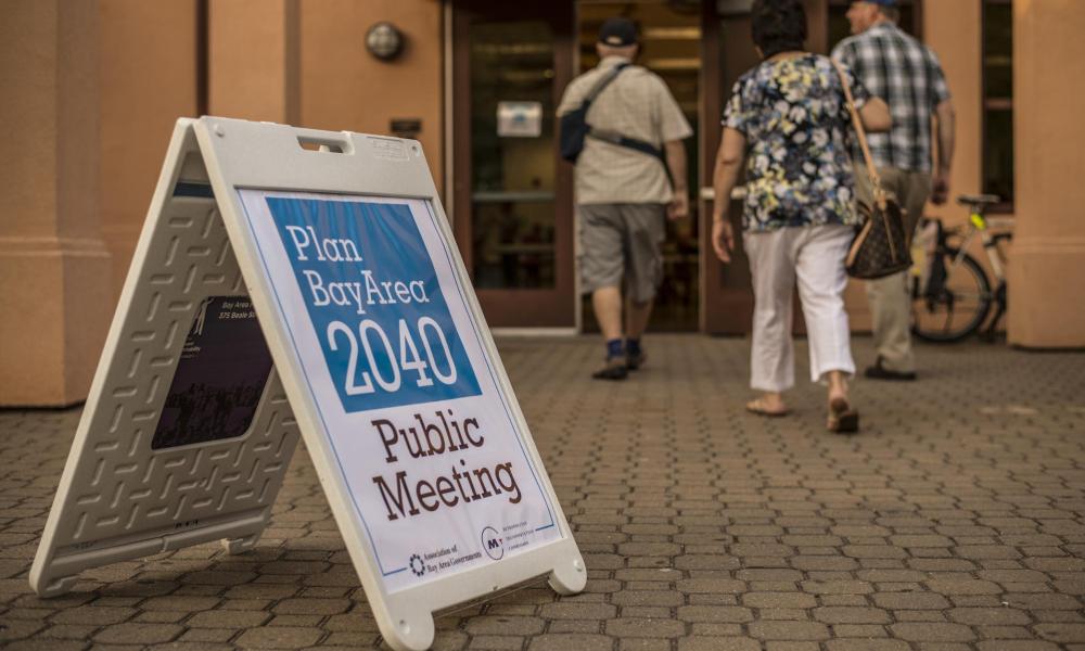 In the foreground a sandwich board advertising the Plan Bay Area 2040 public meeting is displayed, with three adults walking into the building in the background.