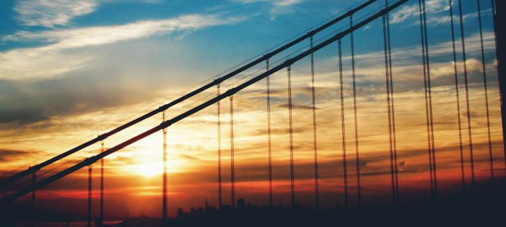 The blue sky transitions to yellow, orange and red as the sun sets behind the suspension cables of the Golden Gate Bridge.